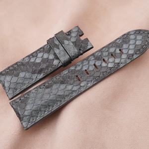Puede incluir: Una correa de reloj de piel de serpiente gris con un patrón texturizado. La correa está hecha de cuero genuino y tiene un cierre de hebilla.