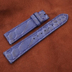 May include: Two blue alligator leather watch straps with stitching. The straps are laid out on a brown suede surface.