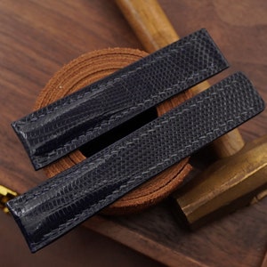 Puede incluir: Dos correas de reloj azul marino con un patrón texturizado similar al de un reptil. Las correas están colocadas sobre una superficie de madera con un rollo de cuero, una herramienta de latón y un mazo de madera en el fondo. Las correas son nuevas.