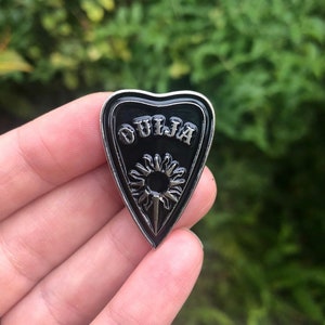 Puede incluir: Pin de metal negro y plateado en forma de corazón con la palabra "OUIJA" escrita en él. El corazón tiene un fondo negro con un diseño de sol plateado en su interior.