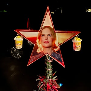 May include: A red star-shaped Christmas tree topper with a picture of a woman inside. The star has two yellow popcorn containers hanging from it.