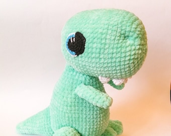 May include: A green crocheted dinosaur plush toy with a blue eye and white teeth. The toy is sitting on a white surface.