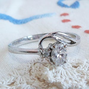 May include: A silver-toned ring featuring a central oval-cut clear stone flanked by two smaller round clear stones. The band has a bypass design that curves around the setting, creating an open, asymmetrical look with a polished metallic finish.