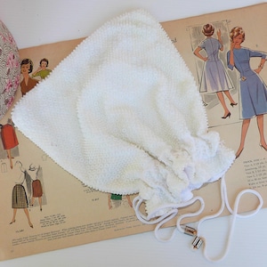 May include: A vintage white beaded drawstring bag with a textured surface and a delicate ruffle detail. The bag has a white cord with decorative rectangular accents. The bag is resting on a vintage sewing pattern.