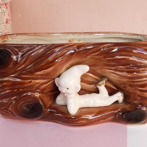 May include: A rectangular brown ceramic planter with a textured wood grain design. A white figurine of a reclining figure with gold accents is inside the planter. The planter has a cream-coloured interior.