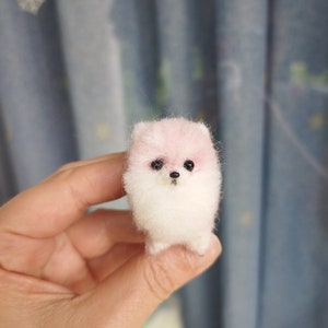 May include: A small, white and pink fluffy dog made of felt. The dog has black eyes and a black nose.