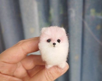 Op de afbeelding: Een kleine, witte en roze pluche hond gemaakt van vilt. De hond heeft zwarte ogen en een zwarte neus.
