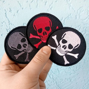 May include: Three embroidered iron-on patches featuring a skull and crossbones design. The patches are circular and have a black background. The skulls are in different colours: red, grey, and white.