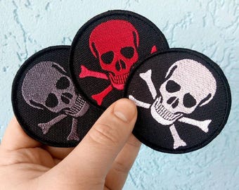 May include: Three embroidered iron-on patches featuring a skull and crossbones design. The patches are circular and have a black background. The skulls are in different colors: red, gray, and white.