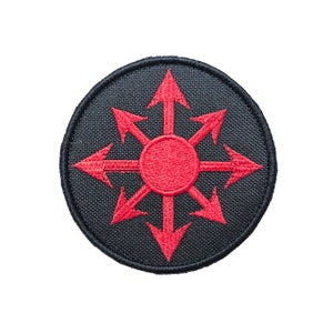 Op de afbeelding: Een zwarte stoffen patch met een rood geborduurd symbool van acht pijlen die in verschillende richtingen wijzen en een stervorm vormen.