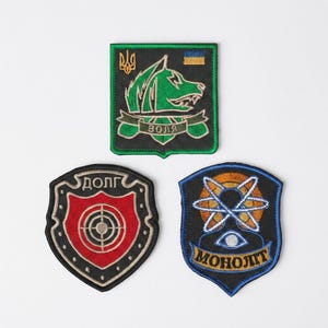 May include: Three embroidered patches featuring distinct designs: a square patch with a green wolf head and the text "ВОЛЯ", a shield-shaped patch with a red target symbol and the text "ДОЛГ", and a shield-shaped patch with an atom and the text "МОНОЛИТ".