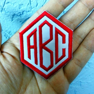May include: A white fabric patch with red embroidered letters "ABC" in a hexagonal shape.