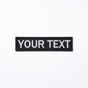May include: A rectangular embroidered patch featuring a black background with a thick black border. The words YOUR TEXT are stitched in white capital letters across the center of the patch, creating a high-contrast design with clear lettering.