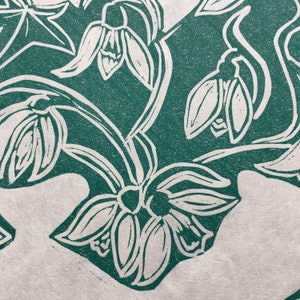 May include: A green and white linocut print of snowdrops. The print features a stylized design of the flowers with their stems and leaves.