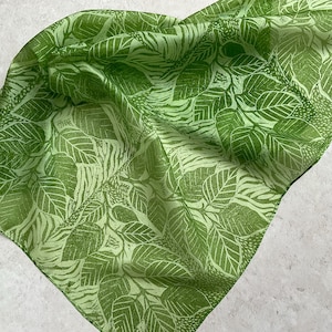 May include: A square, light green scarf with a repeating leaf pattern in a slightly darker shade of green. The leaves have detailed veining, creating a textured appearance. The scarf is folded, showing the pattern from different angles.