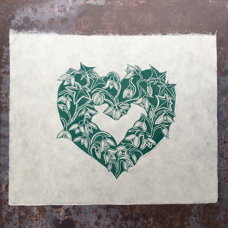 May include: A green linocut print of a heart shape made of snowdrop flowers and leaves. The print is on a white background.