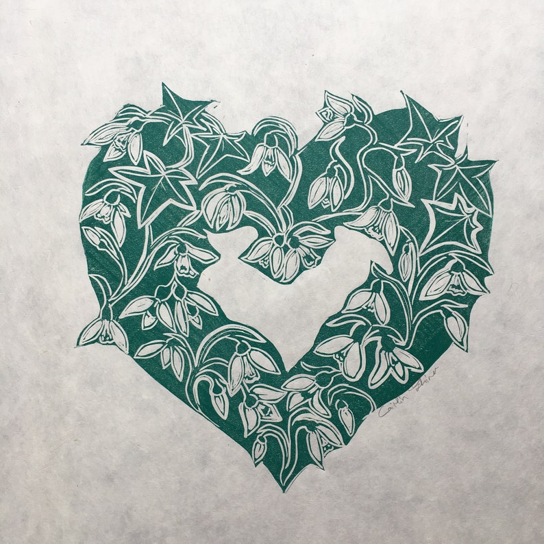May include: A green heart-shaped print featuring a design of white snowdrops and leaves. The print is signed "Caitlin Blair" in the bottom right corner.