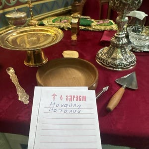 Könnte beinhalten: Eine Sammlung religiöser Gegenstände auf einer roten Tischdecke. Dazu gehören ein silberner Kelch, eine goldene Schale, eine Holzschale, eine Kelle und eine handschriftliche Notiz mit kyrillischem Text. Ein kleines Kreuz und weitere Gegenstände sind ebenfalls vorhanden.