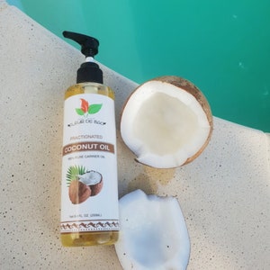Puede incluir: Una botella de vidrio transparente con una tapa de bomba negra y una etiqueta que dice "Fractionated Coconut Oil 100% Pure Carrier Oil" con un logotipo de hoja verde. La botella está sobre una superficie blanca con dos mitades de cáscara de coco.