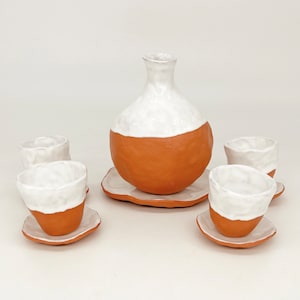 May include: A set of five ceramic cups and a jug, all in a white and terracotta colour scheme. The cups and jug have a textured, uneven surface, giving them a rustic look.