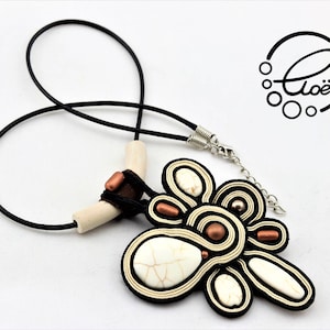Puede incluir: Un collar artesanal con un cordón negro y un colgante con detalles blancos, negros y cobrizos. El colgante tiene un diseño abstracto único con una piedra blanca central y una cadena plateada.