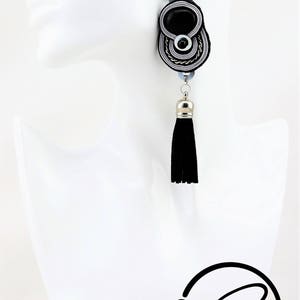May include: Black and silver dangle earring with a black tassel. The earring features a black oval center, surrounded by silver and white accents. A silver-colored metal cap tops the black tassel. The earring is displayed on a white mannequin head.