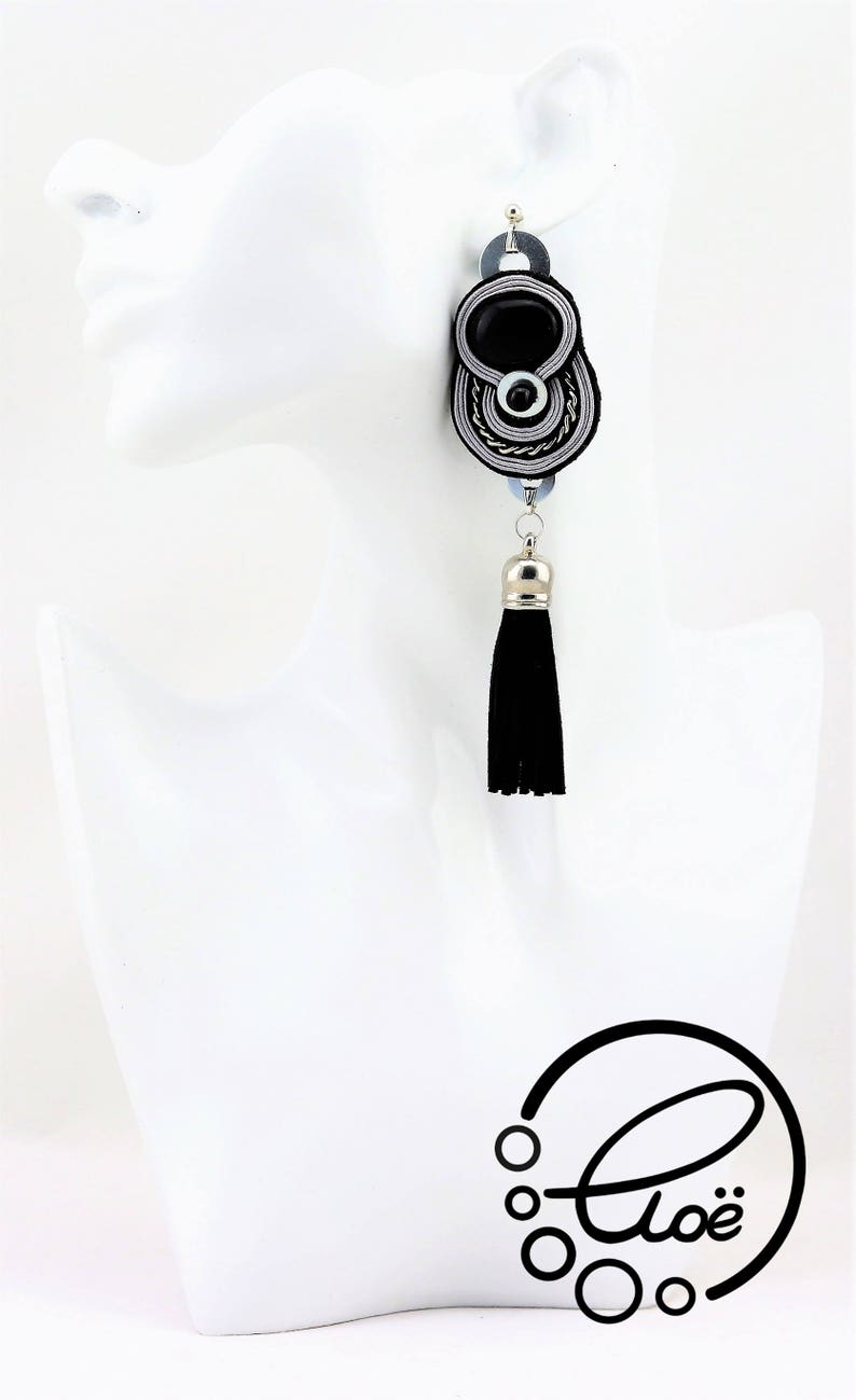 May include: Black and silver dangle earring with a black tassel. The earring features a black oval center, surrounded by silver and white accents. A silver-colored metal cap tops the black tassel. The earring is displayed on a white mannequin head.