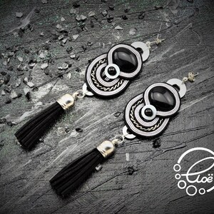 May include: A pair of black and white statement earrings with a unique design. Each earring features a black oval stone, surrounded by intricate white and silver detailing. The earrings have black tassel pendants and silver-toned hardware.