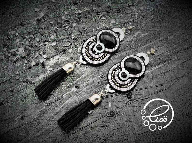 May include: A pair of black and white statement earrings with a unique design. Each earring features a black oval stone, surrounded by intricate white and silver detailing. The earrings have black tassel pendants and silver-toned hardware.