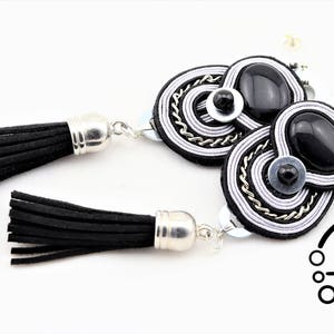 May include: A pair of black and silver tassel earrings. The earrings feature black suede tassels with silver caps. The earring design includes black oval stones, silver chain, and white and silver thread detailing. The earrings are on a white background.