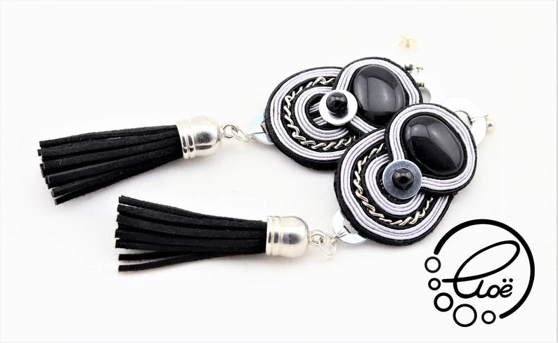 May include: A pair of black and silver tassel earrings. The earrings feature black suede tassels with silver caps. The earring design includes black oval stones, silver chain, and white and silver thread detailing. The earrings are on a white background.