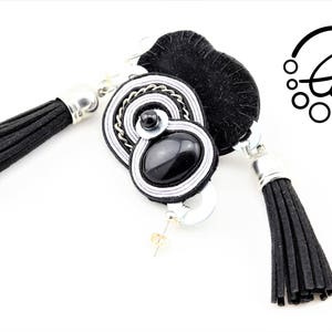 May include: Black and white soutache earrings with a black tassel. The earrings feature a black oval stone and silver-colored metal accents. The earrings have a post back and the logo "Clo&euml;" is visible.