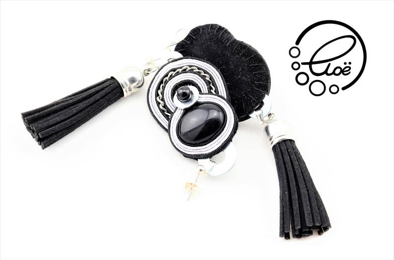 May include: Black and white soutache earrings with a black tassel. The earrings feature a black oval stone and silver-colored metal accents. The earrings have a post back and the logo "Clo&euml;" is visible.