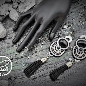 May include: Black and silver-toned earrings with a black oval center stone and a tassel. The earrings have a circular design with silver and white accents. The image also includes a black hand and the logo "Clo&euml;".