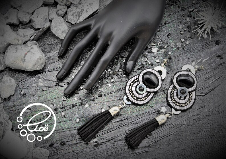 May include: Black and silver-toned earrings with a black oval center stone and a tassel. The earrings have a circular design with silver and white accents. The image also includes a black hand and the logo "Clo&euml;".