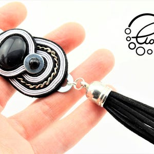 May include: Black and silver-toned statement bracelet with a black tassel. The bracelet features a central design with a black stone, silver and black accents, and a chain detail. The logo "Clo&euml;" is visible.