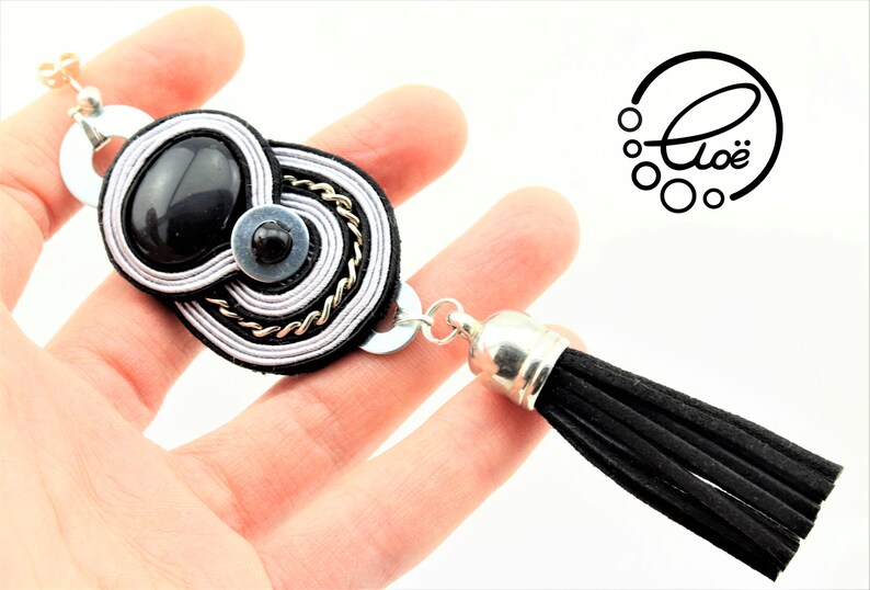 May include: Black and silver-toned statement bracelet with a black tassel. The bracelet features a central design with a black stone, silver and black accents, and a chain detail. The logo "Clo&euml;" is visible.