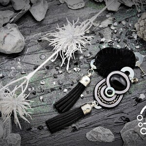 May include: Black and silver tassel earrings with a black and white pendant. The earrings are displayed on a dark, textured surface with decorative white floral accents and small stones. The logo "Clo&eacute;" is visible in the bottom right corner.