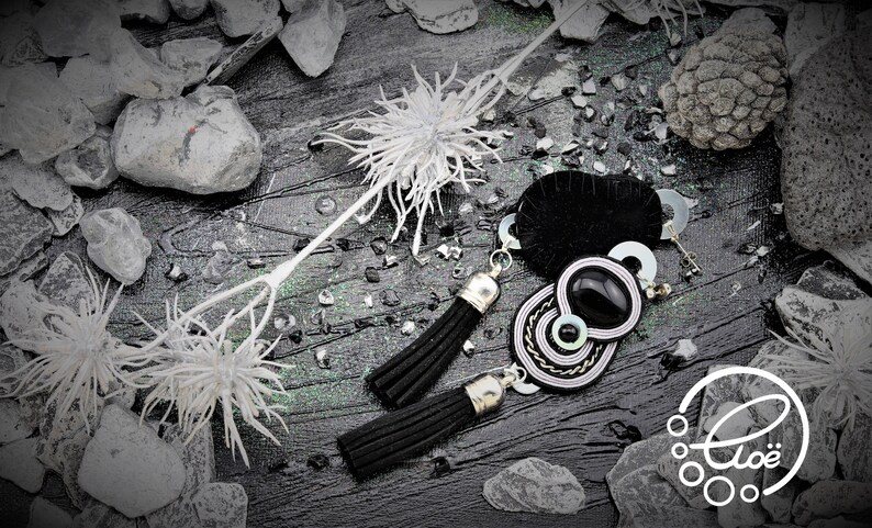 May include: Black and silver tassel earrings with a black and white pendant. The earrings are displayed on a dark, textured surface with decorative white floral accents and small stones. The logo "Clo&eacute;" is visible in the bottom right corner.