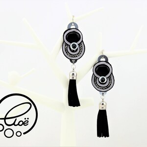 May include: A pair of black and silver dangle earrings with a black tassel. The earrings have a central black oval stone, surrounded by silver and black woven detailing. The earrings are displayed on a white decorative stand.