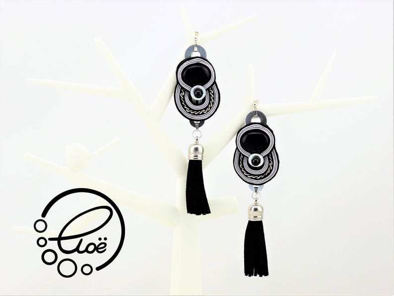 May include: A pair of black and silver dangle earrings with a black tassel. The earrings have a central black oval stone, surrounded by silver and black woven detailing. The earrings are displayed on a white decorative stand.