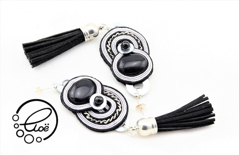 May include: A pair of black and silver tassel earrings. Each earring features a black oval stone, surrounded by silver and white thread, with a silver chain accent. The earrings have black tassels and silver-toned metal findings.