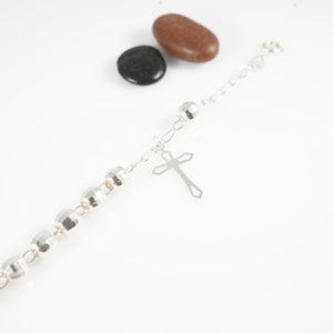 Puede incluir: Un brazalete de plata con un dije de cruz y cuentas redondas. El brazalete está sobre una superficie blanca.