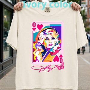 Puede incluir: Una camiseta de manga corta color marfil con un gráfico de carta de juego que muestra un colorido retrato pop-art de una mujer. El diseño incluye la letra Q, símbolos de corazones y el texto Dolly en cursiva con una mariposa rosa debajo.