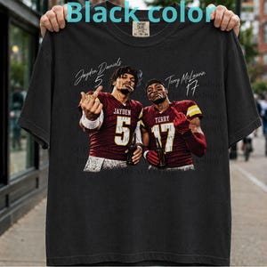 May include: A black t-shirt featuring a graphic print of two football players in maroon jerseys with the names Jayden Daniels 5 and Terry McLaurin 17. Both players are shown holding bottles and making hand gestures. The shirt has a crew neck design.