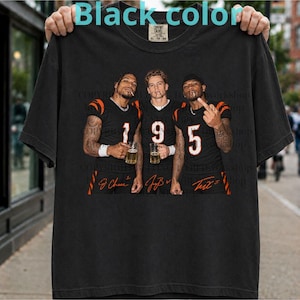 May include: A black short-sleeve t-shirt featuring a graphic print of three football players in black jerseys with orange numbers 1, 9, and 5. Each player holds a glass of beer. The graphic includes orange signatures below each player.