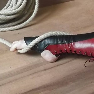 Puede incluir: Un guante de cuero bicolor en negro y burdeos con un diseño de cordones en el lateral y una abertura para el pulgar. El guante se muestra con una cuerda gruesa y trenzada de color natural que pasa por la abertura del pulgar.
