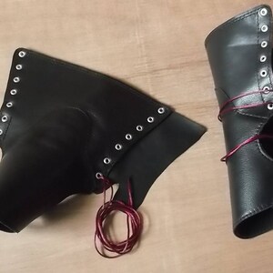May include: A pair of black leather-like arm guards or greaves featuring silver-toned metal eyelets along the edges. The pieces are laced with thin, dark red cord, allowing for adjustable closure. The material has a smooth, matte finish.