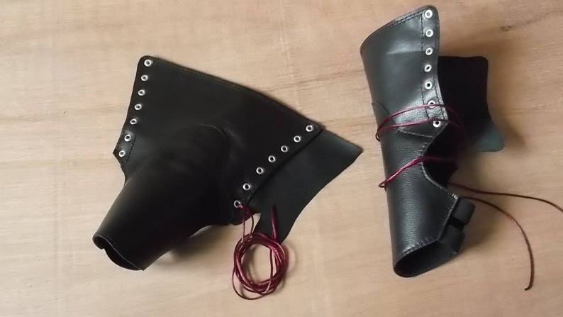 May include: A pair of black leather-like arm guards or greaves featuring silver-toned metal eyelets along the edges. The pieces are laced with thin, dark red cord, allowing for adjustable closure. The material has a smooth, matte finish.