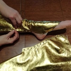 Peut inclure: Une paire de jambi&egrave;res en tissu dor&eacute; m&eacute;tallis&eacute; dot&eacute;es d'une s&eacute;rie d'&oelig;illets m&eacute;talliques argent&eacute;s sur les bords lat&eacute;raux. Le tissu pr&eacute;sente une surface textur&eacute;e et r&eacute;fl&eacute;chissante, con&ccedil;ue pour envelopper le bas de la jambe avec un la&ccedil;age.